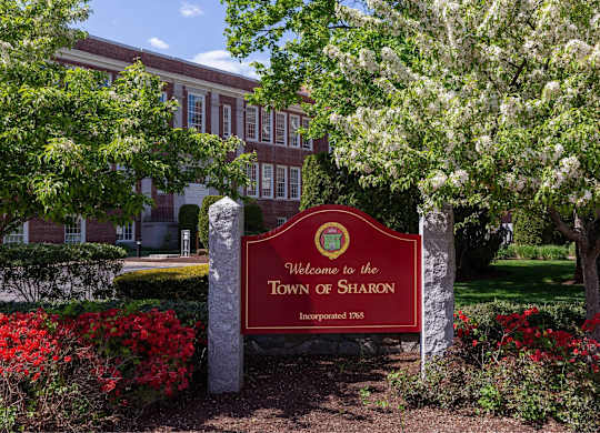 A sign welcoming people to the Town of Sharon stands in front of a building.