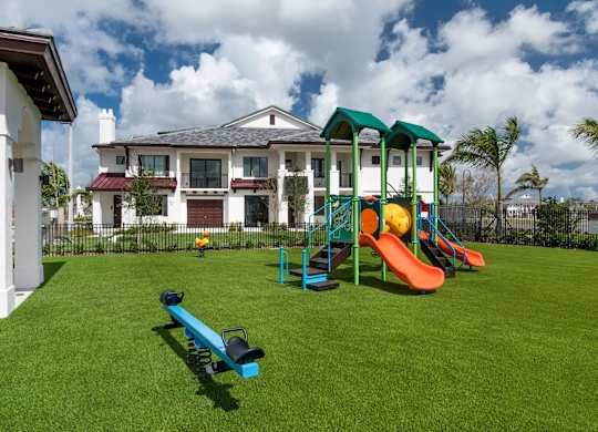 A playground with a slide, swings and a seesaw in front of a large house.