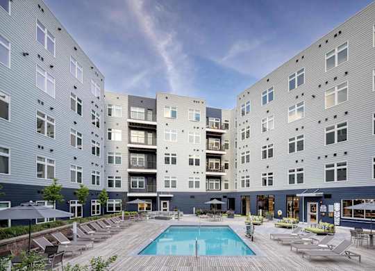 A large swimming pool is surrounded by lounge chairs in the courtyard of a modern apartment complex.