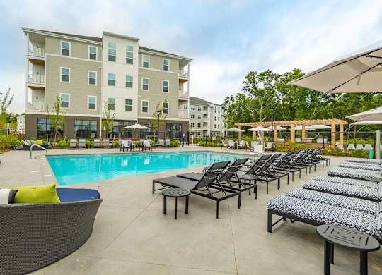 A large swimming pool surrounded by lounge chairs and umbrellas in front of a multi-story apartment building.