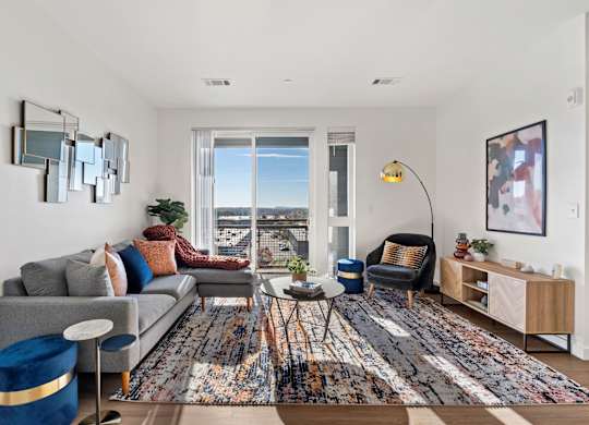 A living room with a grey couch, a blue ottoman, a wooden side table, and a large window with a view of the city.