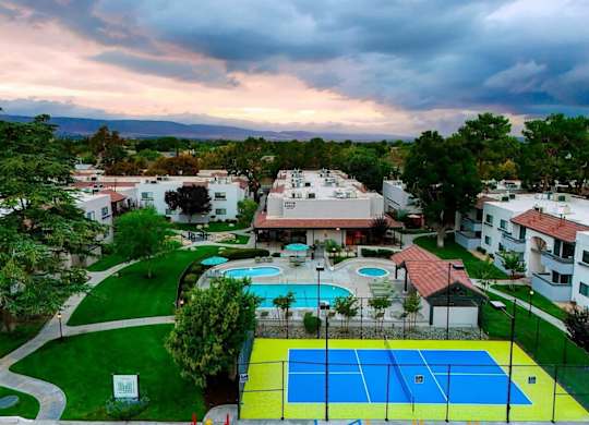 A tennis court is surrounded by a pool and apartment buildings.