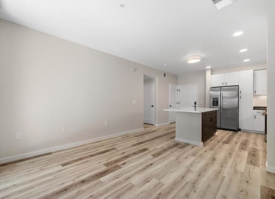 an empty living room with a kitchen and a stainless steel refrigerator