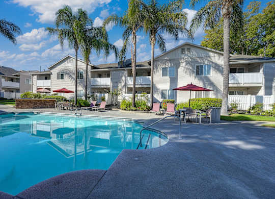 Community swimming pool with large palm trees in the background as well as one of the community apartment buildings. There are tables and chairs around the pool deck at The Legacy Apartments, Antelope