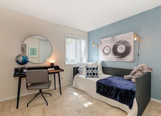 a bedroom with a daybed along the wall and a desk with mirror on the wall.  Bedroom also has carpet and window.