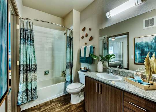 Apartment guest bathroom with white tub and blue shower curtain. Toilet can be seen next to a modern wood vanity and large mirror at Craft at Gilbert and Baseline Apartments, Mesa