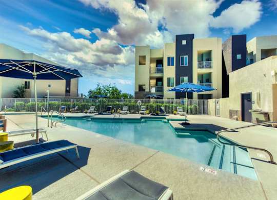 Pool with buildings in the background and patio umbrella up front. Four lounge chairs line the pool.