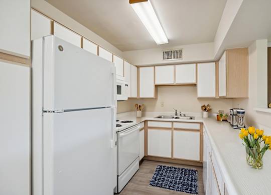 A white refrigerator stands in a kitchen with wooden cabinets and a sink.