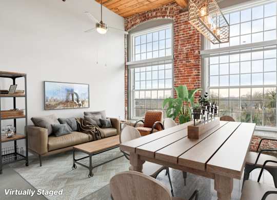 Virtually staged living room in Converse Mill Lofts in Spartanburg, SC with high ceilings, exposed beams, brick walls, large windows and a kitchen with stainless steel appliances.