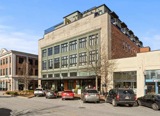 Exterior of Aug Smith on Main, a factory converted to apartments. Cars are parked outside of it. The building is next to the other shops that line the street.