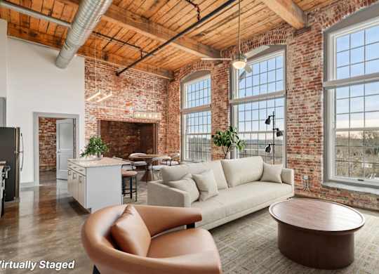 Staged living room in Converse Mill Lofts in Spartanburg, SC with high ceilings, exposed beams, brick walls, large windows and a kitchen with stainless steel appliances.