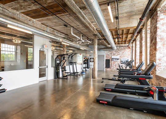 Loft Apartments for Rent in Spartanburg, SC - Converse Mill Lofts - Fitness Center with High Ceiling, Exposed Ductwork and Brick Walls, Large Windows, Large Mirror, Free Weights, and Cardio Equipment.