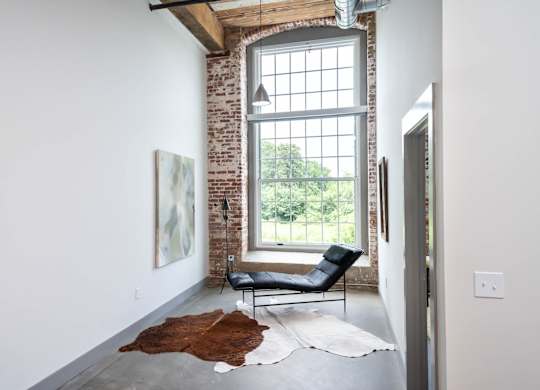 Loft Apartments Near Downtown Spartanburg, SC - Converse Mill Lofts - Living Room with High Ceiling, Large Window, Chaise Lounge, Pendant Lighting, Couch, Coffee Table, Armchairs, and Paintings.