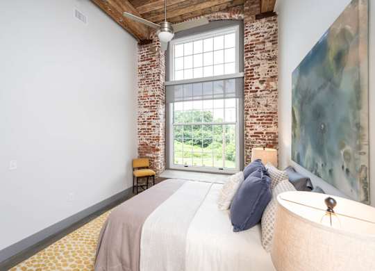 Loft Studios Spartanburg, SC - Converse Mill Lofts - Bedroom with High Ceiling, Concrete Flooring, Large Bed, Large Painting, Nightstands, Lamps, Chair, Large Windows, and Exposed Brick Walls.