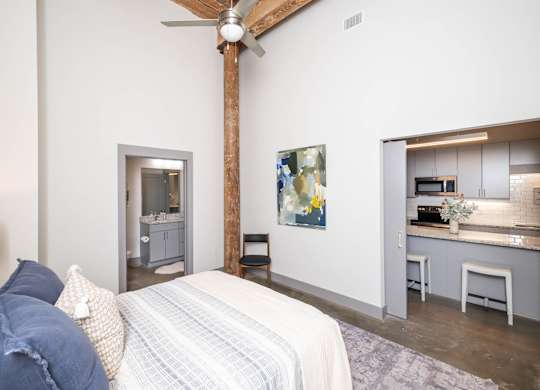 Loft Studios Spartanburg, SC - Converse Mill Lofts - Bedroom with High Ceiling, Concrete Flooring, Large Bed, Large Painting, Nightstands, Lamps, Chair, and bathroom.
