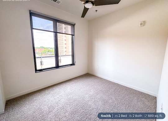 A second bedroom with a carpeted floor, a ceiling fan, and a large window overlooking a cityscape.