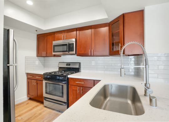 Apartment kitchen with stainless steel appliances, white tile back splash, white counter tops, and cherry wood cabinets.