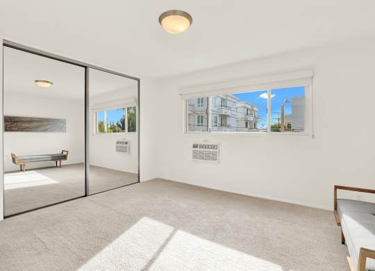 A white room with a bed and a window showing a view of a house at Wooster Encore Apartments, Los Angeles, California