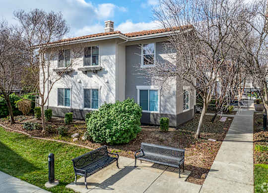 A house with a grey and white exterior is surrounded by a well-kept lawn and a sidewalk with two benches at Cornerstone at Gale Ranch Apartments, California, 94582