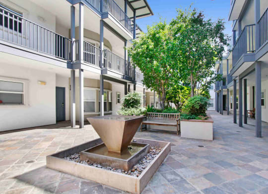 a fountain sits in the middle of a courtyard with trees and benches at Toluca Court, Toluca Lake