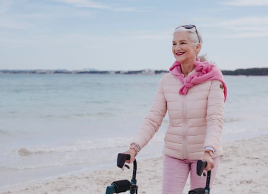 A woman in a pink jacket and pants is pushing a walker on a beach.
