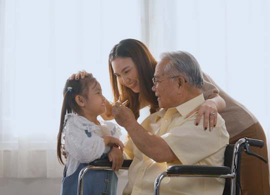 A woman is giving a young girl a haircut while an elderly man sits in a wheelchair nearby.