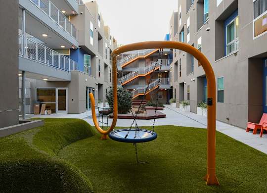 A playground with a swing set in the middle of a grassy area in front of apartment buildings.