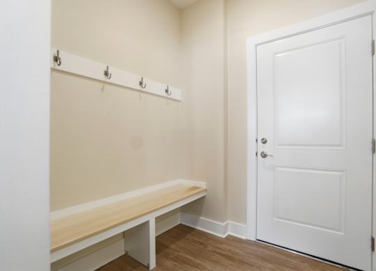 A white door is on the right side of a room with a bench and coat hooks on the wall.