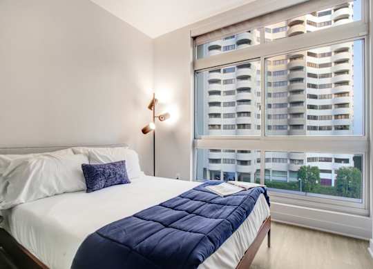 A bedroom with a bed, a window with a view of a building, and a wall lamp.at Wilshire Margot, Los Angeles, 90024 