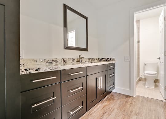 Long single vanity area with granite countertops, wood inspired flooring, dark brown cabinets, bathroom on the right