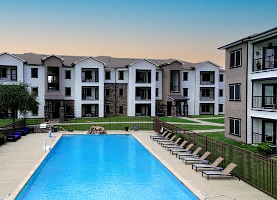 A large swimming pool is surrounded by lounge chairs in front of apartment buildings.