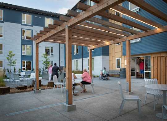 A wooden pergola is over a patio with people sitting and a motorcycle parked outside a building.