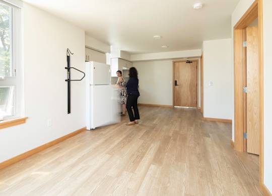 A woman is standing in a room with wooden floors and white walls.