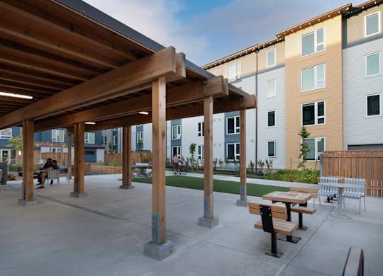 A wooden pergola is over a concrete patio with benches and picnic tables.