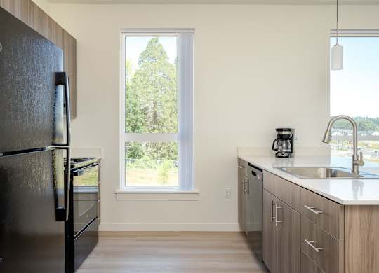 A black fridge stands in a kitchen with a window showing trees outside.