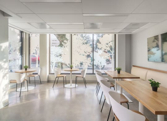 a room with wooden tables and white chairs with large windows in the background
