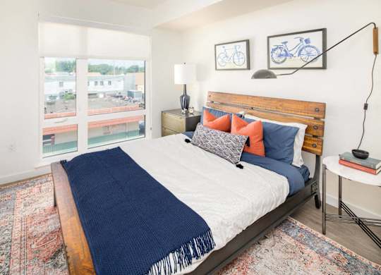 A bedroom with a bed, a nightstand, and a window overlooking a parking lot.