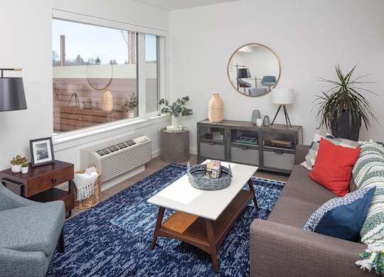 A living room with a grey couch, a blue rug, and a round mirror on the wall.
