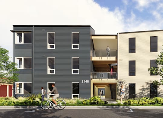 A person riding a bicycle in front of a grey building with the number 3048 on it.