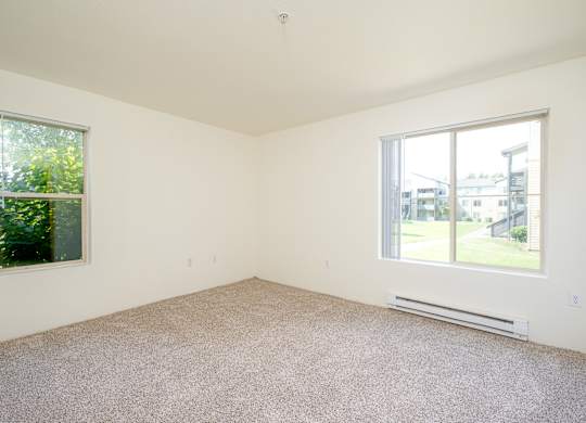A room with a carpet floor, a window with a view of a building and trees outside, and a wall-mounted air conditioner.
