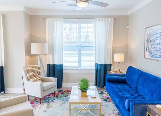 A living room with a blue couch, a white chair, and a small table with a plant on it. at Foxwood Apartments, Raleigh-Durham