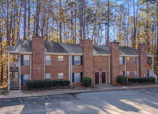 A red brick building with a black roof and chimneys is surrounded by trees.