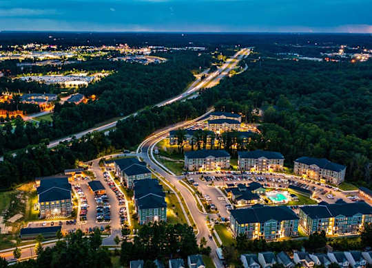 A nighttime aerial view of a resort with a lit swimming pool and cars parked in the parking lot at Foxwood Apartments, Raleigh-Durham, NC, 27616