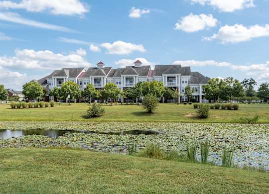 A large white house with a red roof is surrounded by a grassy lawn and a pond with lily pads.