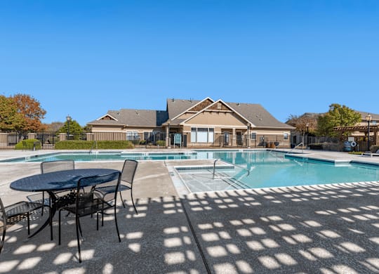 Swimming pool with large tanning deck, lounge chairs, round table and chairs with clubhouse exterior in background and native landscape