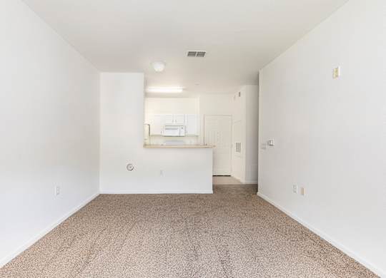 A white room with a carpeted floor and a small kitchenette in the corner.
