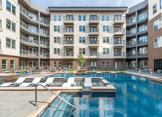 Pool courtyard with pool in the middle and chairs around pool with view of apartment building and balconies and tanning chairs