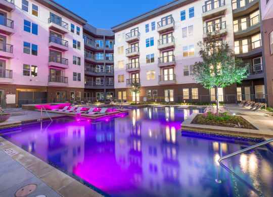 Courtyard pool area during night time with purple lights in water and view of surrounding apartments and balconies with tree and flower bed next to pool