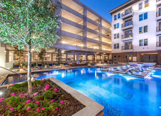 Courtyard pool area during night time with bright lights in water and view of surrounding apartments and balconies and parking garage behind pool with tree and flower bed next to pool