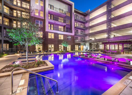 Courtyard pool area during nighttime with purple lights in water and view of surrounding apartments and balconies with parking garage in background